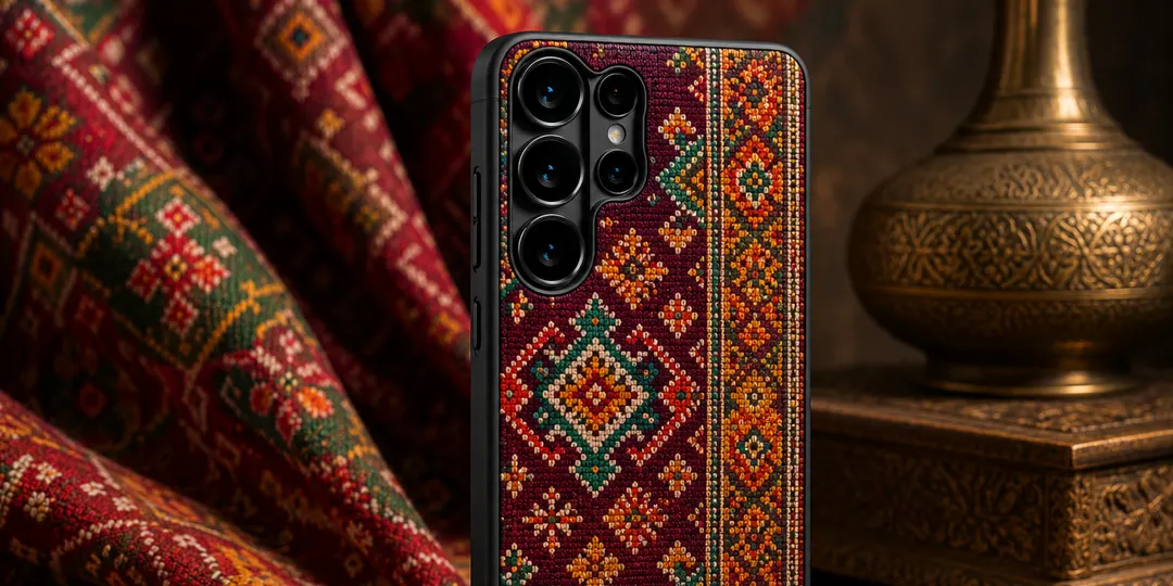 PITAKA phone case with a woven kilim pattern in maroon, green, and gold, styled beside draped textile and a brass vessel
